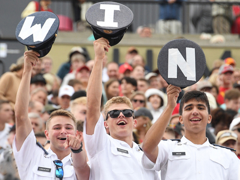 ϲ cadets in stands cheer a sports team and hold up hats that spell out W I N